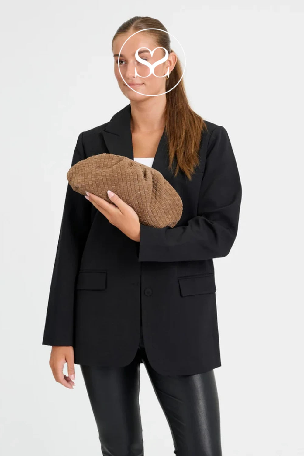 A woman wearing a black blazer and black leather-look pants holds a brown textured crocodile-pattern clutch bag against a plain white studio background.