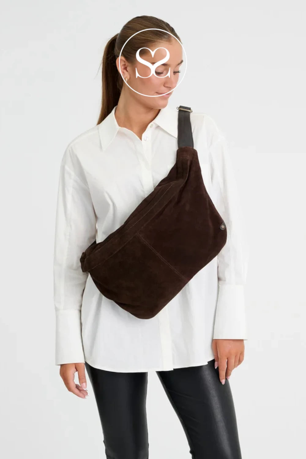 A woman wearing a white button-up shirt and black pants models a dark brown suede crossbody bag with a black strap against a light gray background.