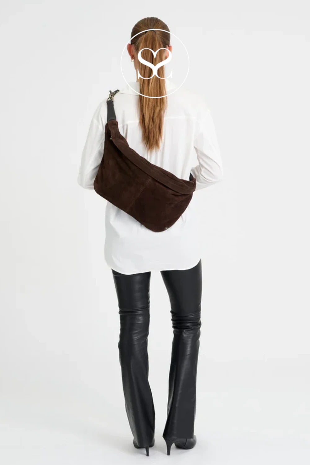 A woman shown from the back wearing a dark brown suede crossbody bag over a white shirt and black flared leather pants, against a plain white studio background.