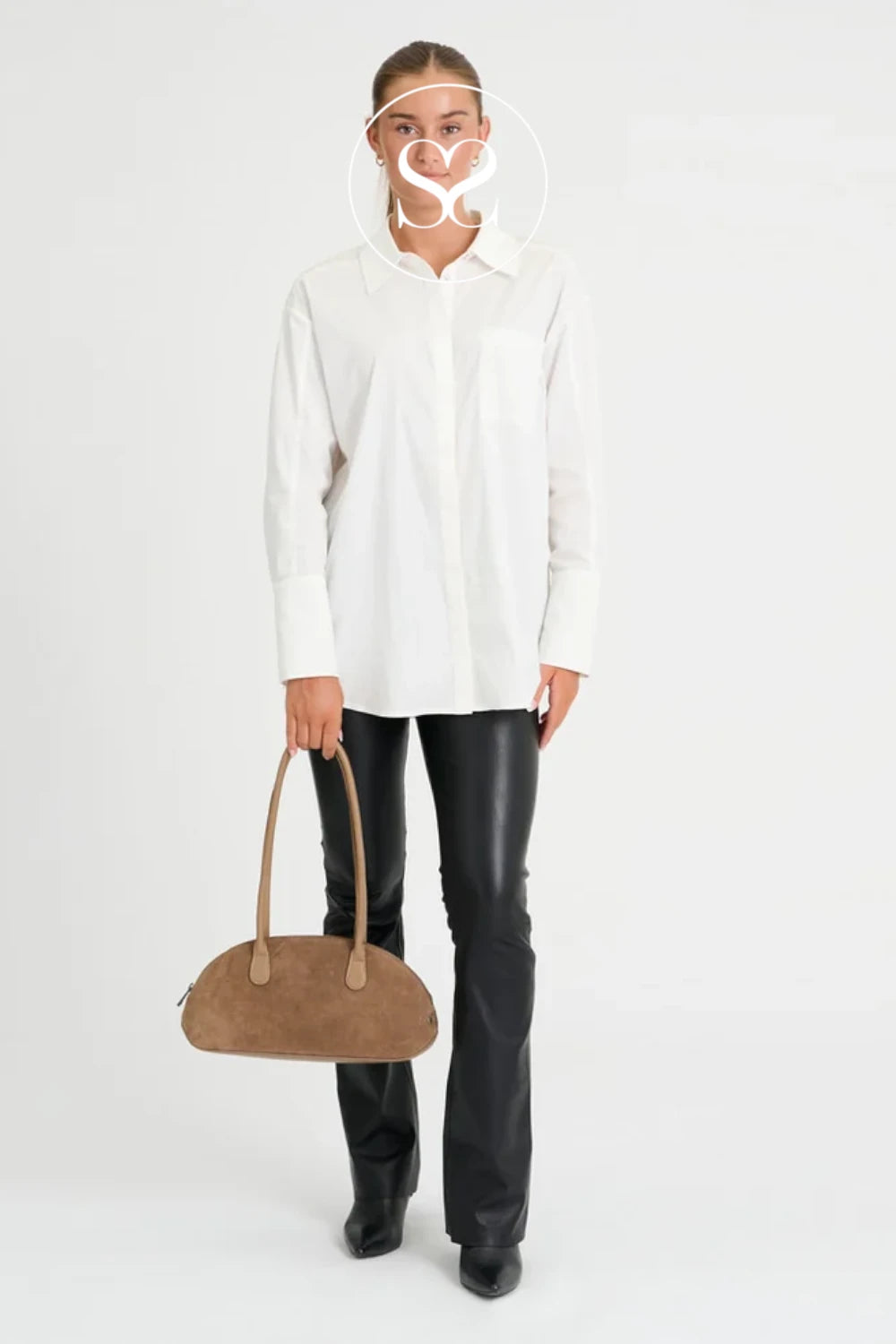 A model stands against a light gray studio background wearing an oversized white button-up shirt, black leather pants, and black boots, while holding a taupe-brown suede handbag. A white circular monogram logo overlay is centered on the model's face.