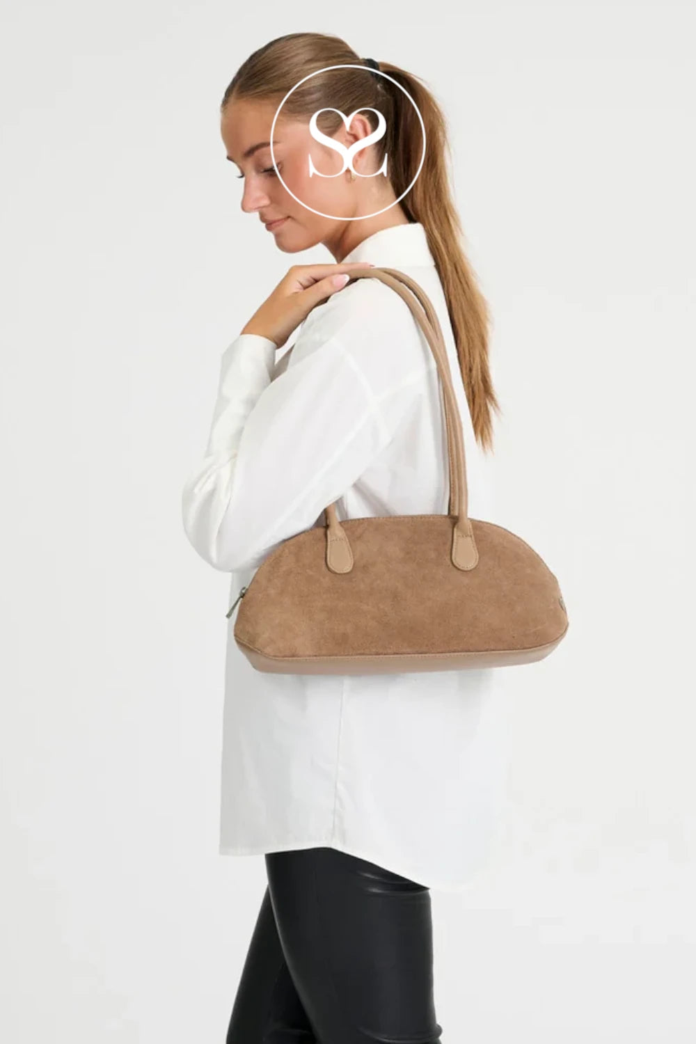 A woman in a white blouse and black pants carries a taupe suede dome handbag with matching shoulder straps against a plain white studio background.