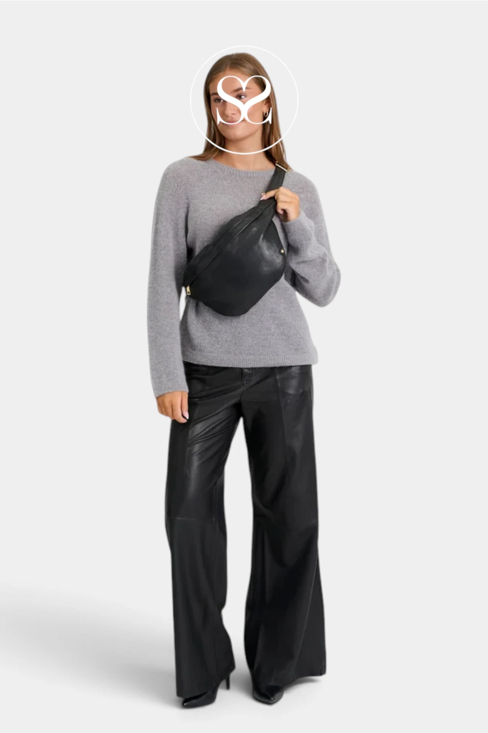A woman modeling a black crossbody sling bag, wearing a gray knit sweater and black wide-leg leather pants, against a light gray studio background.