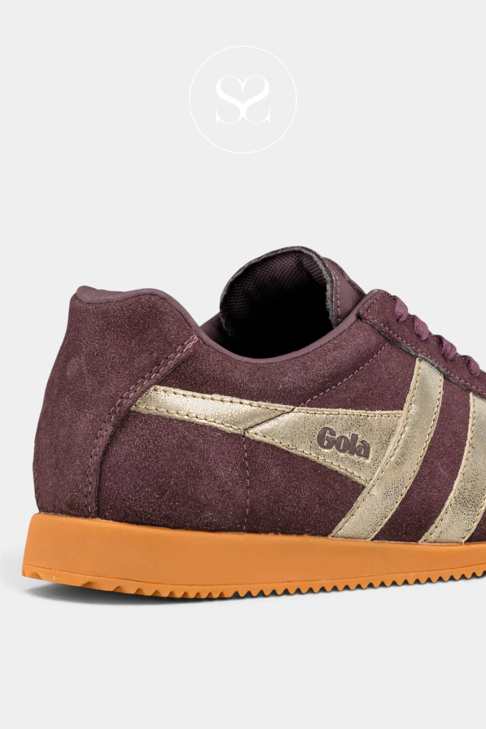 A close-up side view of a low-top sneaker in dark burgundy suede with metallic gold side stripes and a gum orange sole, shown on a light gray background.