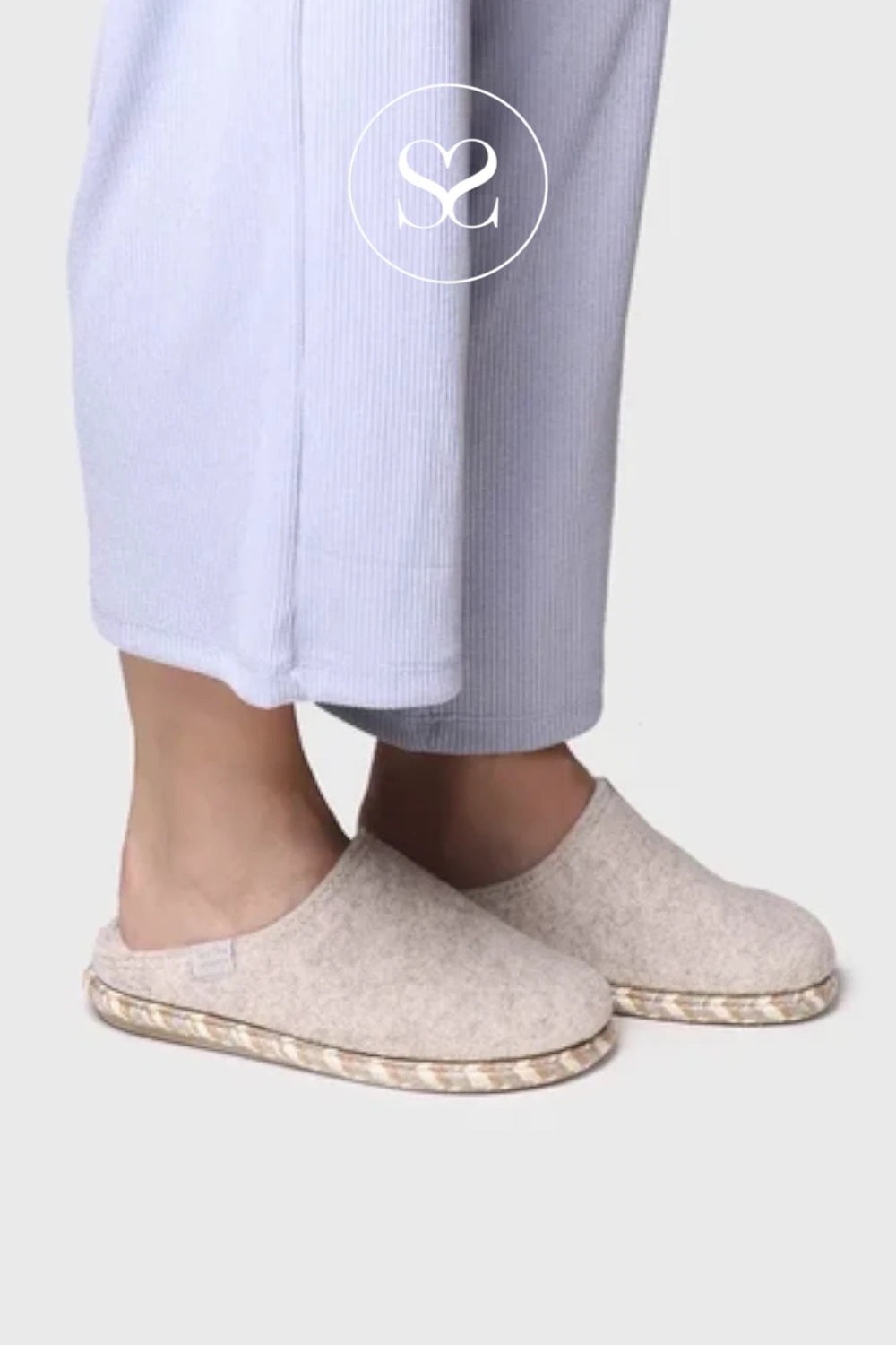 A person wearing light beige slip-on slippers with braided jute-style soles, paired with pale lavender ribbed pajama pants, shown against a light gray background.