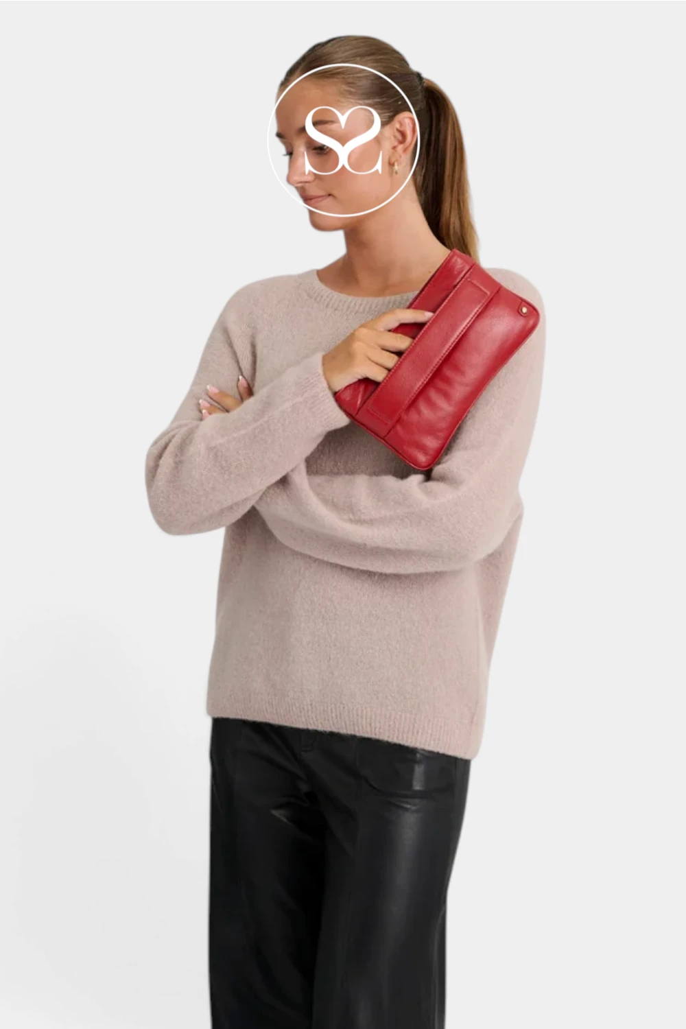 A woman wearing a fuzzy light beige sweater and black pants holds a red leather clutch bag against her arm, photographed against a plain light gray studio background.