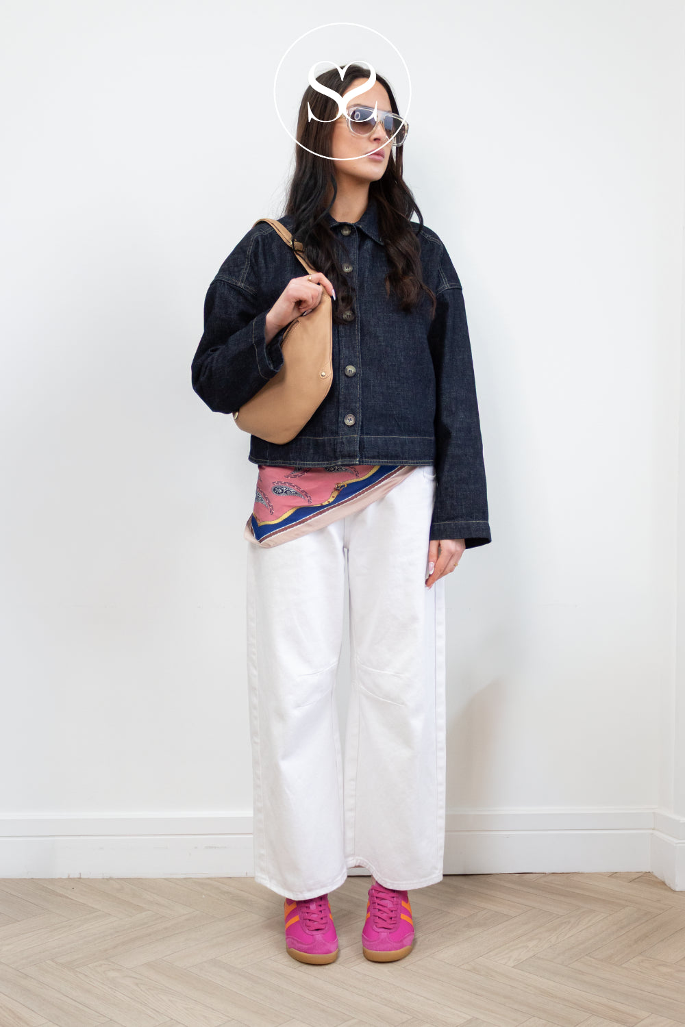 Woman wearing a denim jacket, white pants, and pink gola trainers against a white wall.