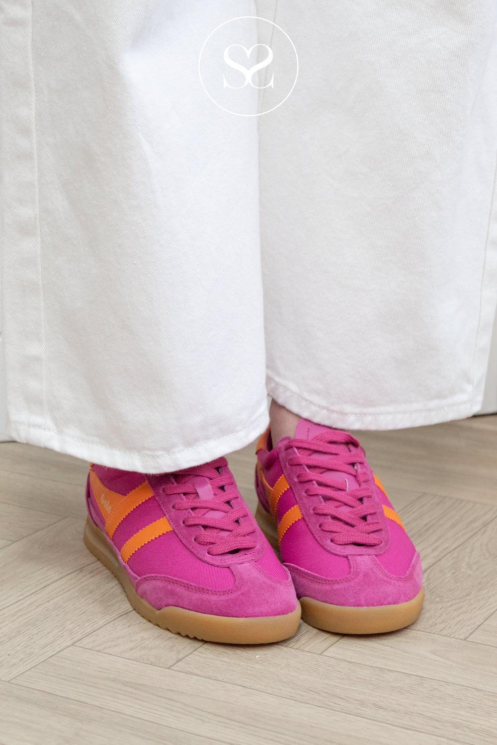 Pink sneakers with orange accents from Gola worn with white pants on a wooden floor.