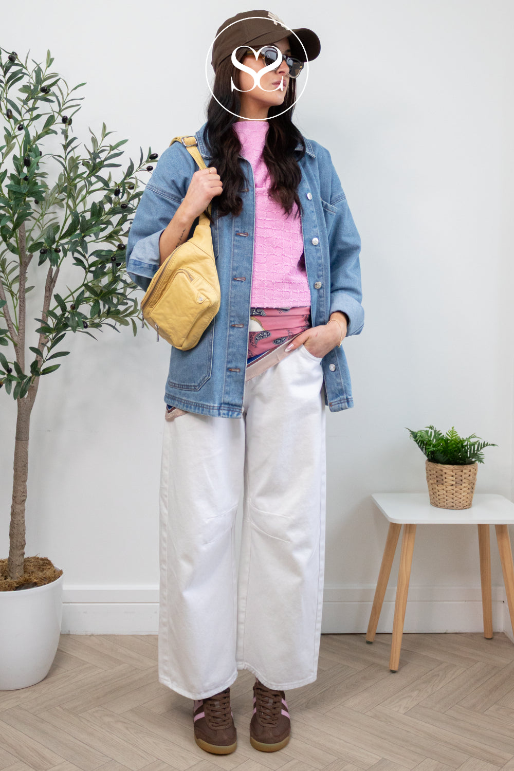 Person wearing a denim jacket, pink shirt, white pants, and brown Gola trainers in a room with plants and a small table.