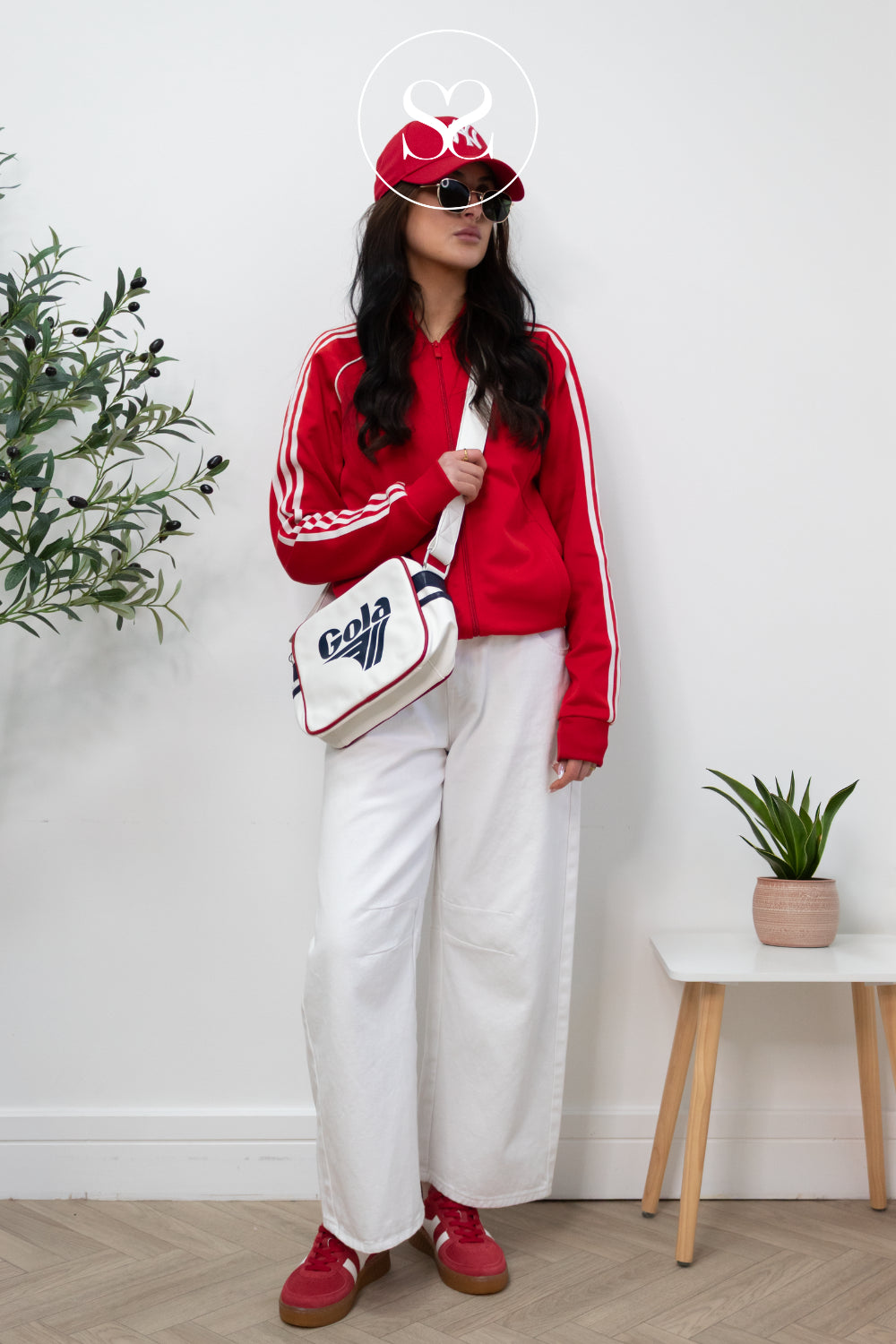 Person wearing a red tracksuit with white stripes, white pants, and red gola trainers, holding a white and red gola messenger bag indoors.