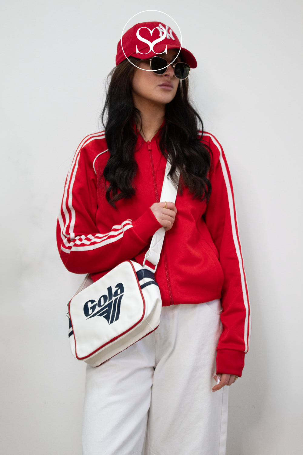 Person wearing a red tracksuit with white stripes, holding a white gola bag with branding, against a plain background.