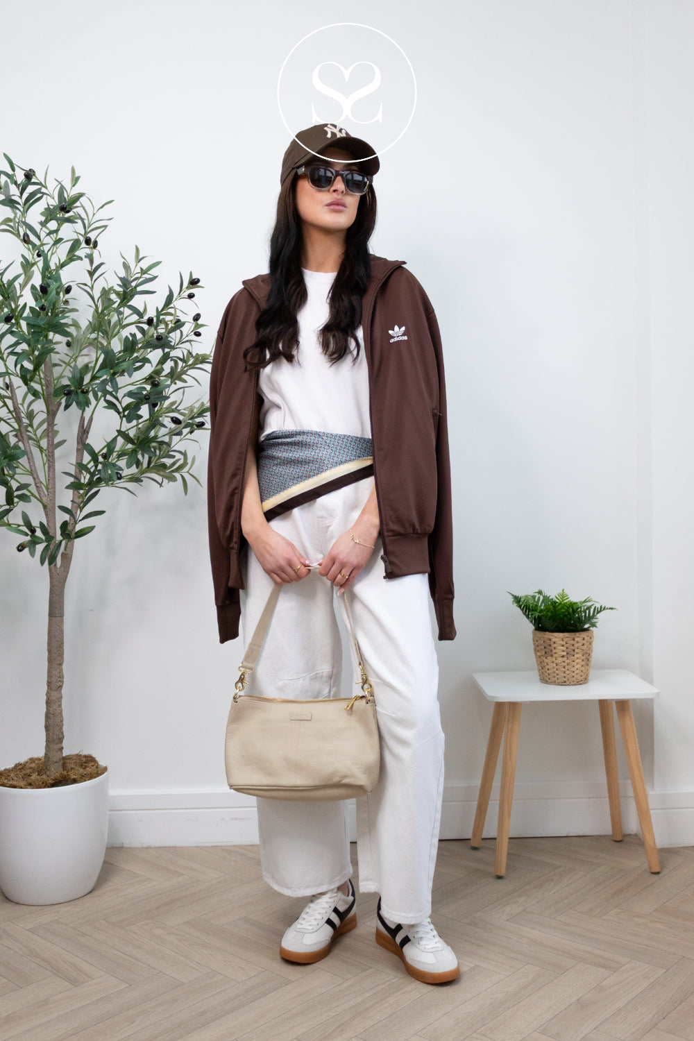 Woman wearing a brown jacket and white pants with Gola Retro White & Black sneakers Cyclone standing indoors with a plant and small table in the background.