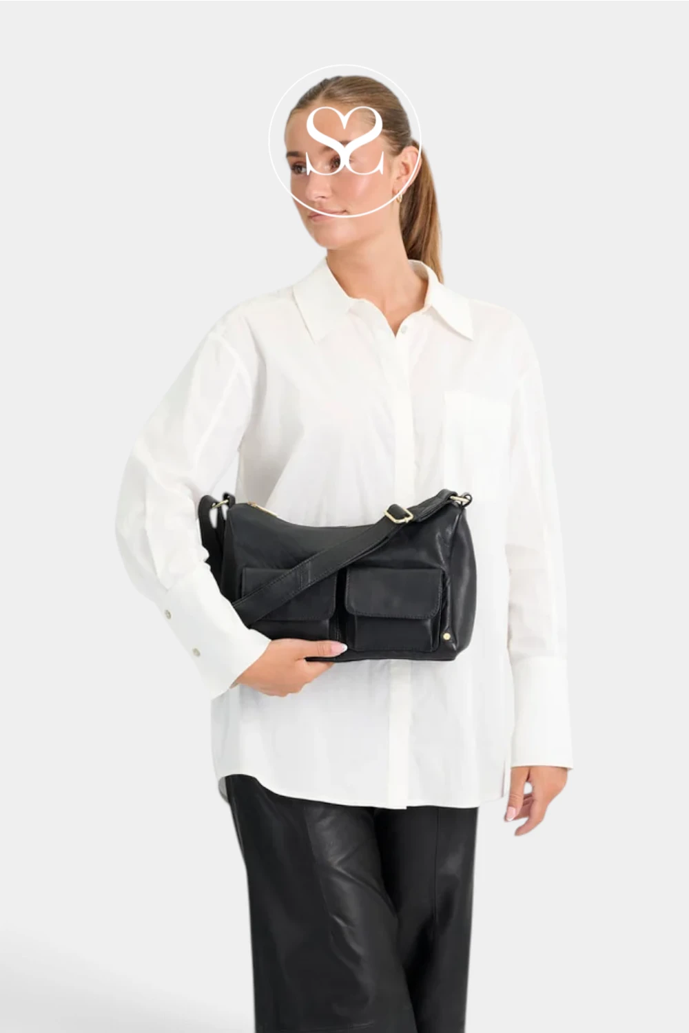 A model wearing a white button-down shirt and black pants carries a black leather shoulder bag with gold-tone hardware against a light gray studio background.
