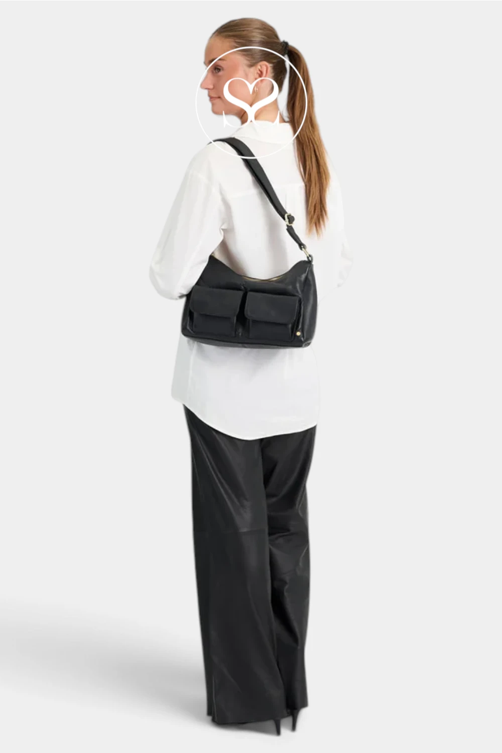 A woman shown from the back wearing a black shoulder bag with multiple front pockets, styled with a white long-sleeve top and black wide-leg pants against a light gray studio background.