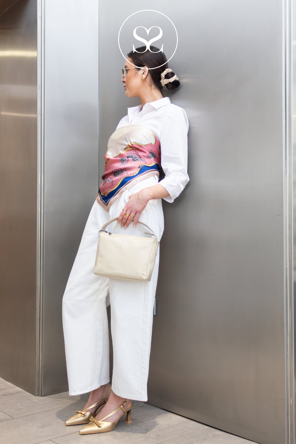 Woman in workwear outfir with a modern touch, leaning against a metallic wall showcasing gold high heels from Hispanitas