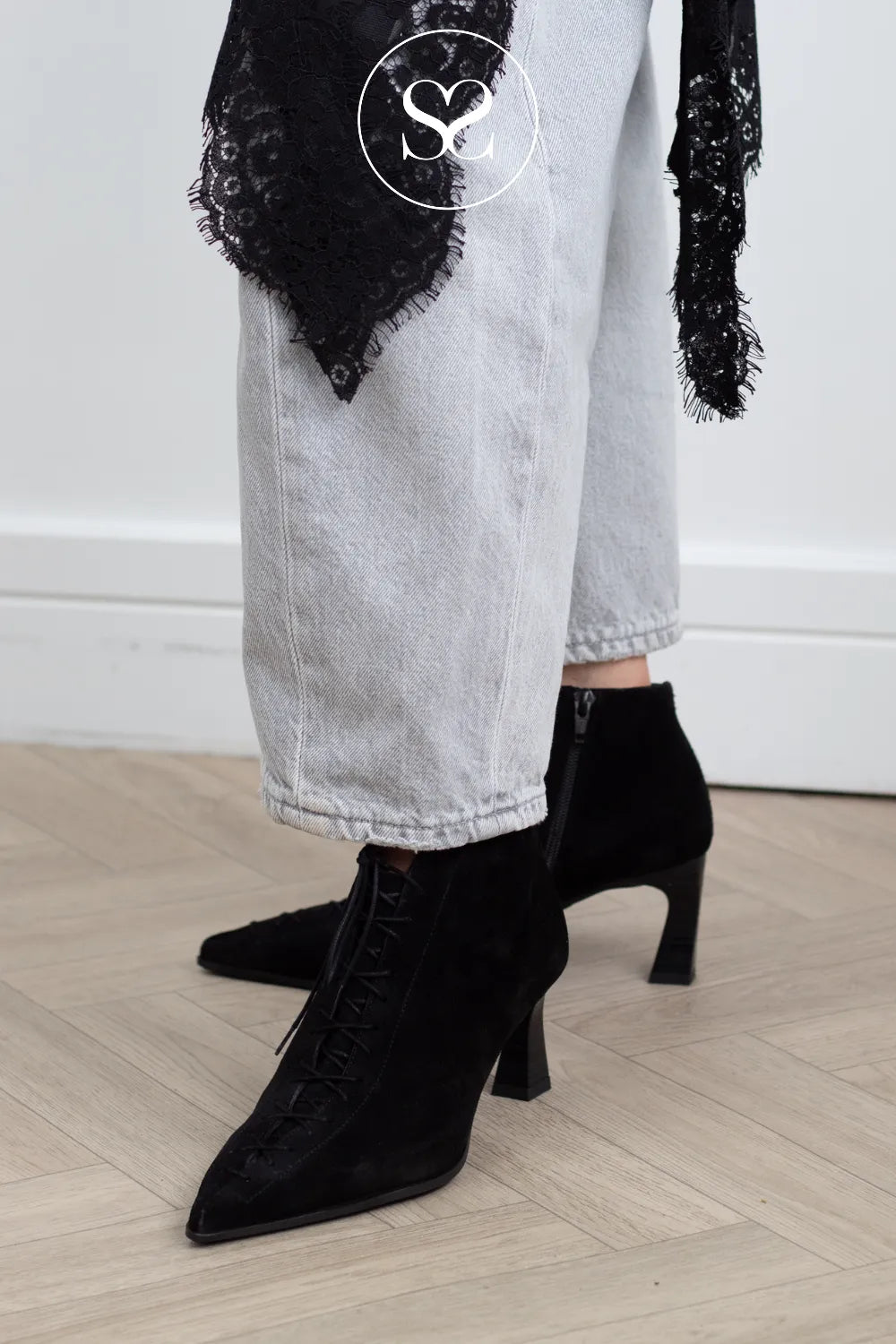 A person wearing black pointed-toe ankle boots with a kitten heel, styled with light-wash wide-leg jeans and a black lace wrap, standing on a light wood floor.