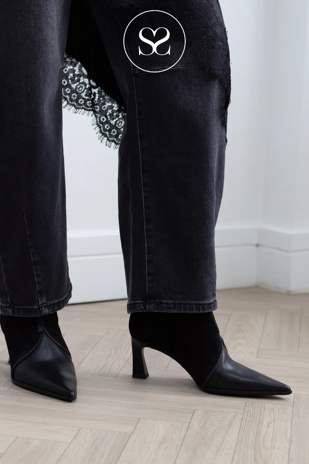 Black pointed-toe heeled ankle boots worn with dark washed jeans, shown from the knees down in an indoor room with a light wood floor and white wall.