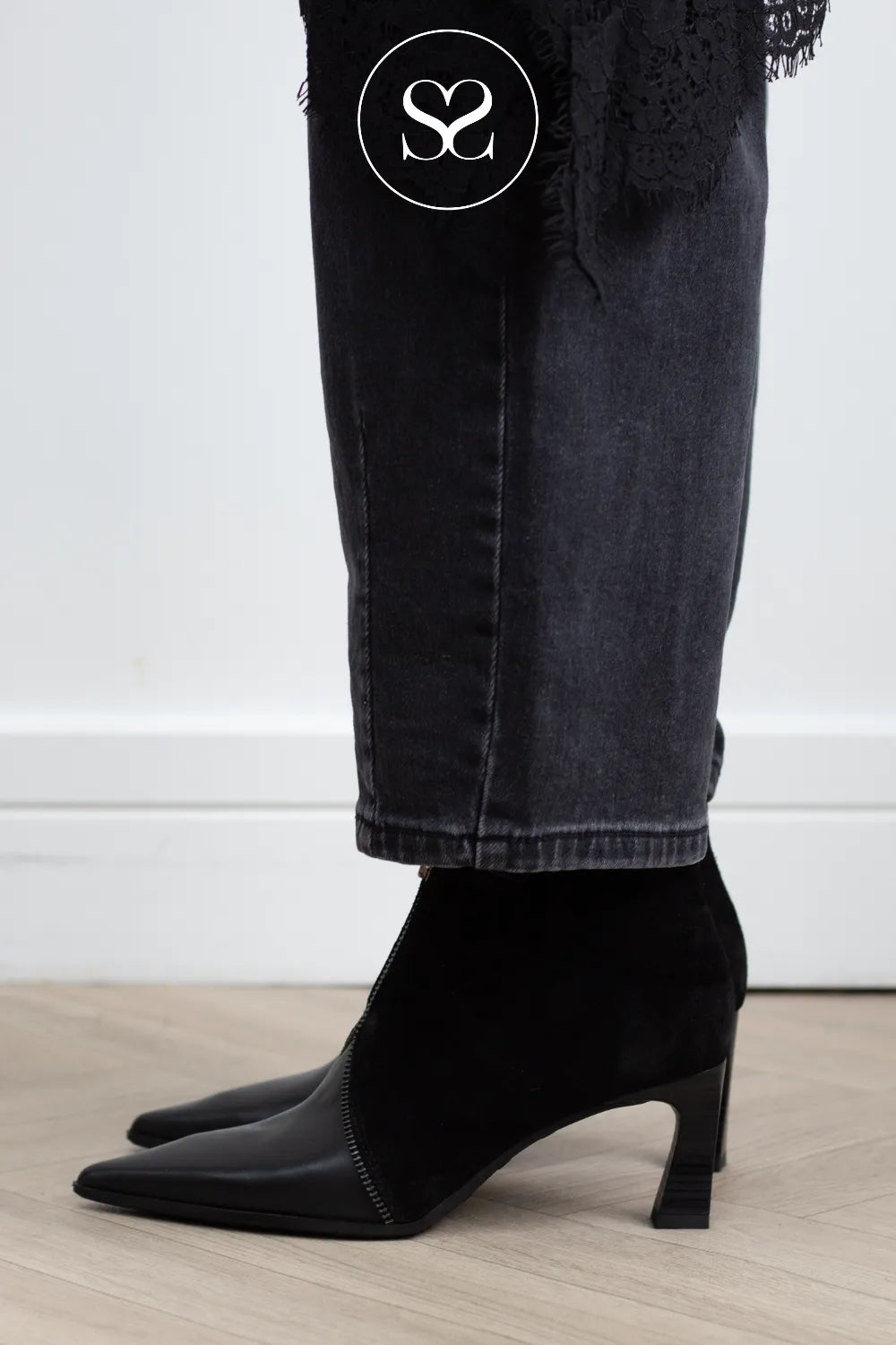 Black pointed-toe ankle boots with a velvet finish and a low block heel, shown on a model wearing dark gray jeans. A white circular logo overlay is visible at the top.