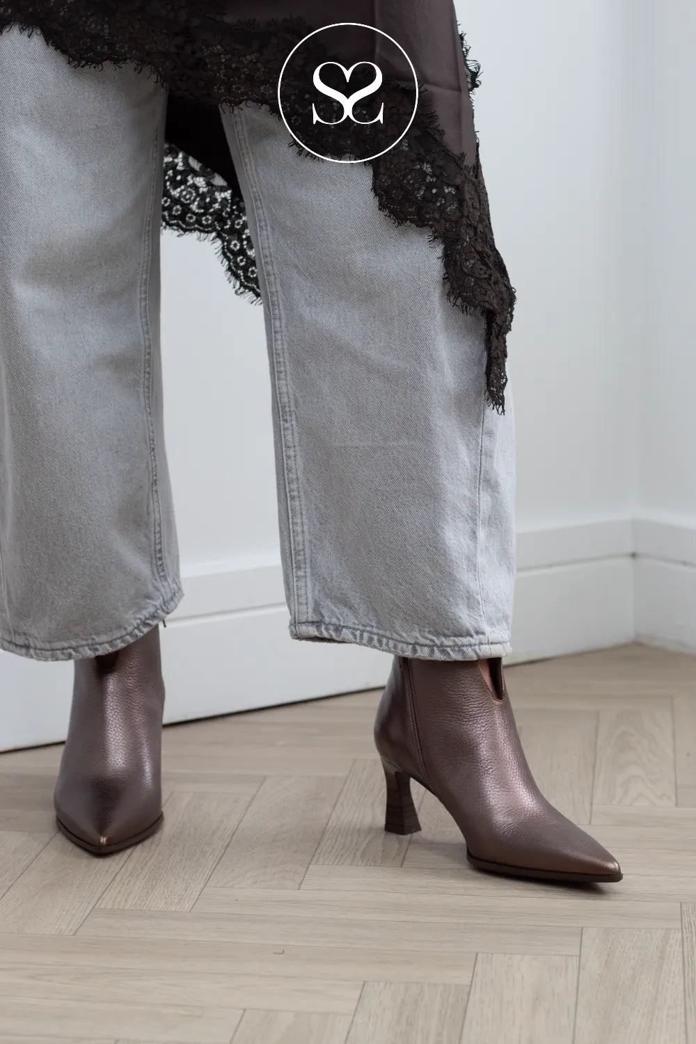 A person wearing pointed-toe heeled ankle boots in a dark brown leather, paired with light gray jeans and a black lace-trim top, standing on a light wood herringbone floor.