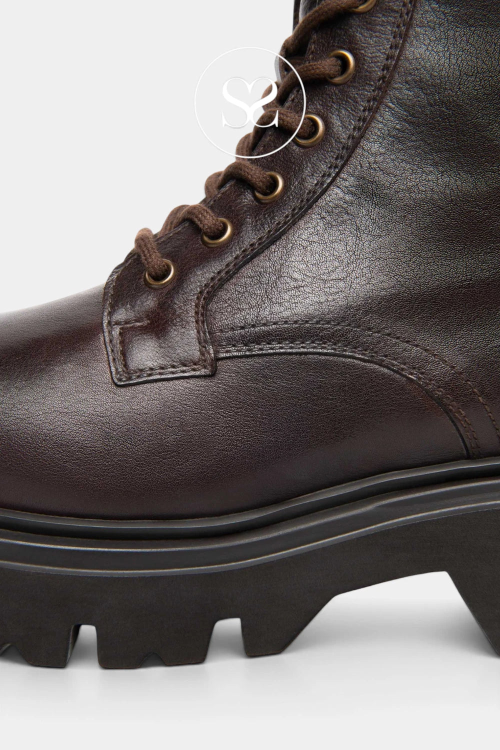 Close-up of a dark brown leather lace-up boot with gold eyelets and a chunky black lug sole, shown against a light gray background.