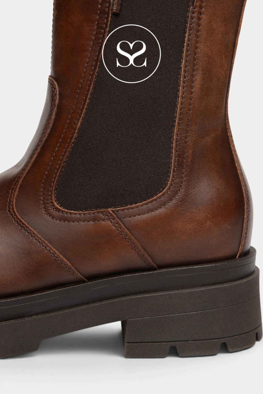 Close-up of a brown leather Chelsea boot with a dark elastic side panel and a white circular monogram logo near the top.
