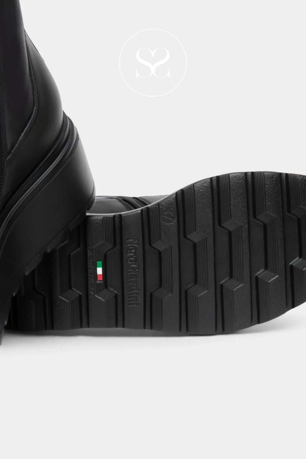 Close-up of the black sole of a leather boot, showing the tread pattern and a small green, white, and red brand tab, on a light gray background.