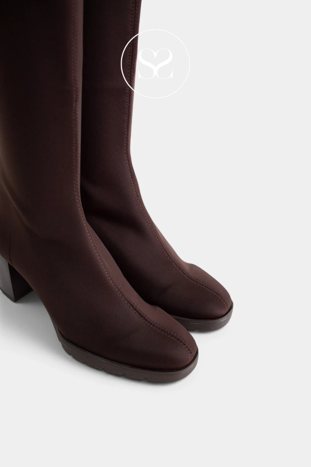 A close-up of a pair of dark brown knee-high boots with a matte finish, featuring a low block heel and square toes, set against a light gray background.