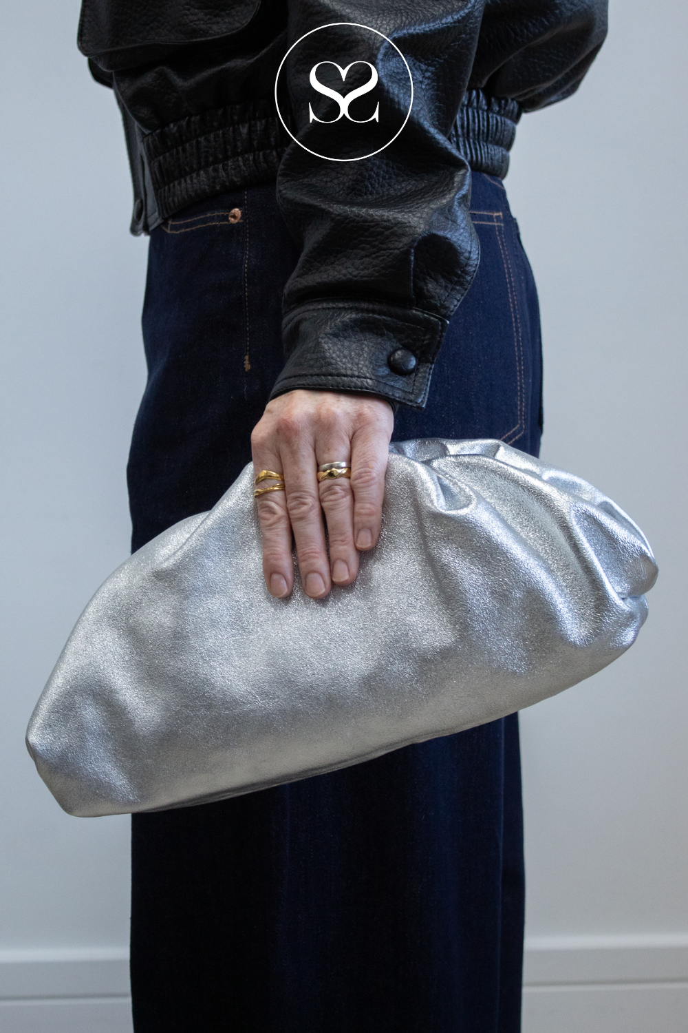 A person wearing a black leather jacket and dark blue jeans holds a metallic silver crinkled clutch bag. The person is wearing gold rings, and there is a circular 'SS' logo watermark on the jacket.