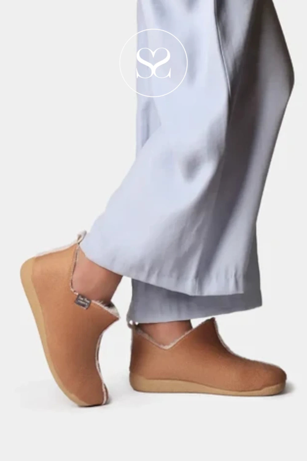 A person wearing light gray wide-leg trousers and tan slip-on clogs with a small side zipper, shown against a plain light gray studio background. A white circular 'SC' logo watermark is visible near the top.