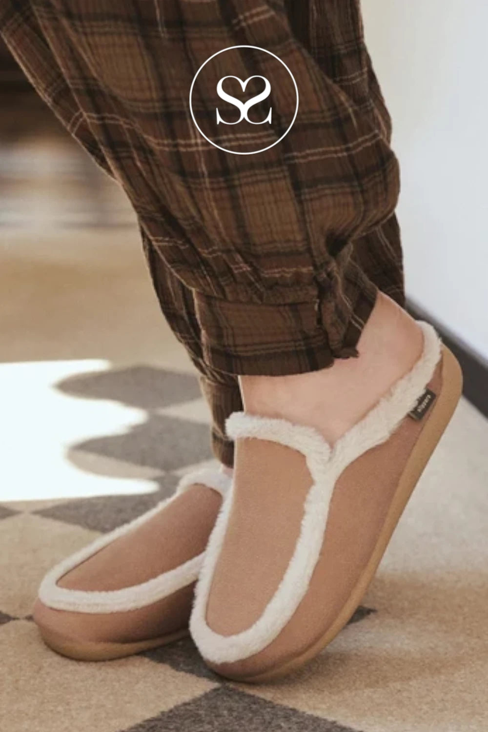 A person wearing tan slip-on slippers with white faux-fur trim and brown plaid pajama pants, standing on a patterned rug indoors. A small black tag is visible on the side of one slipper, and a circular 'SC' logo watermark appears near the upper legs.