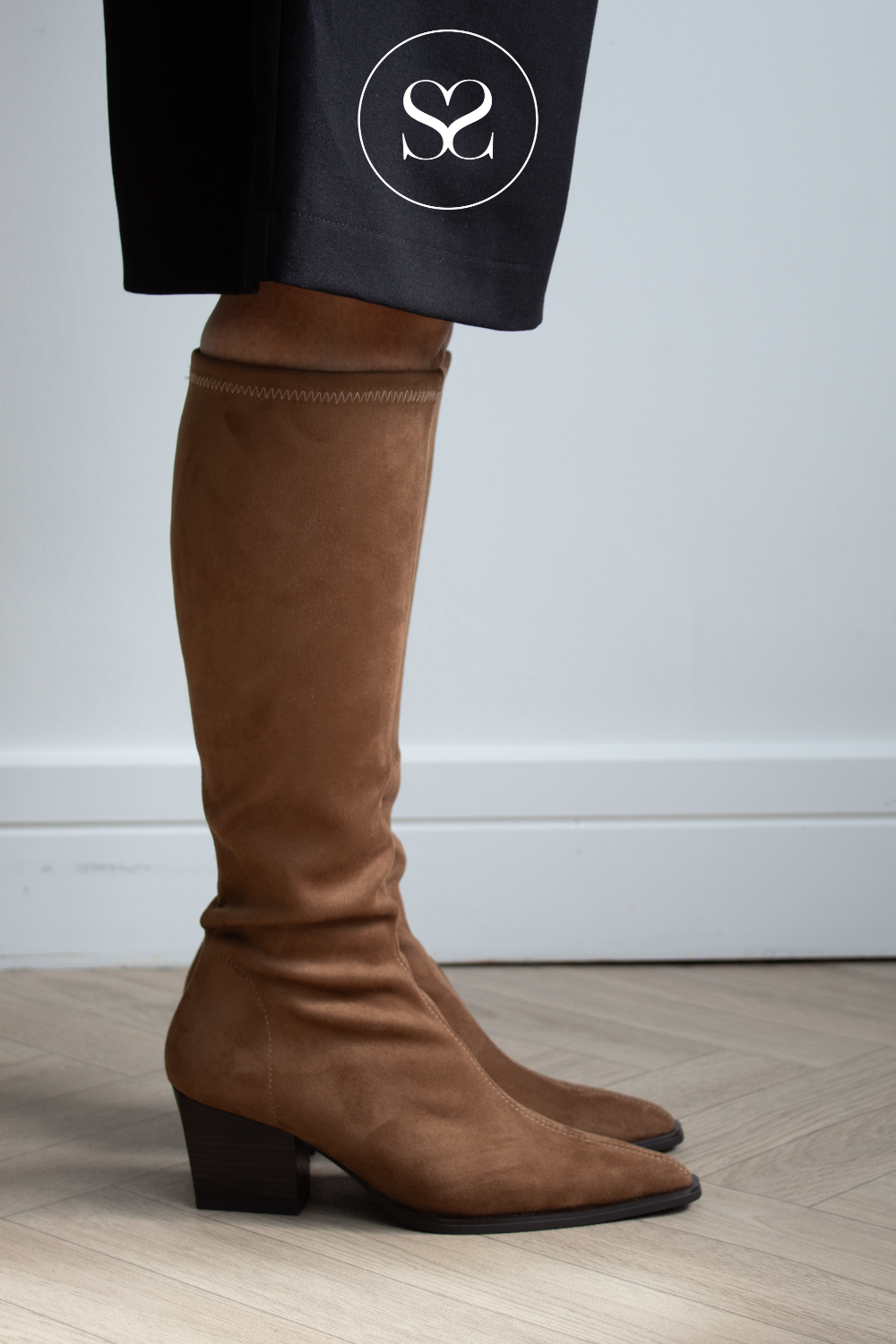 A pair of brown suede knee-high boots with low block heels, shown on a model wearing black trousers. The background is a clean white wall and light wood floor.