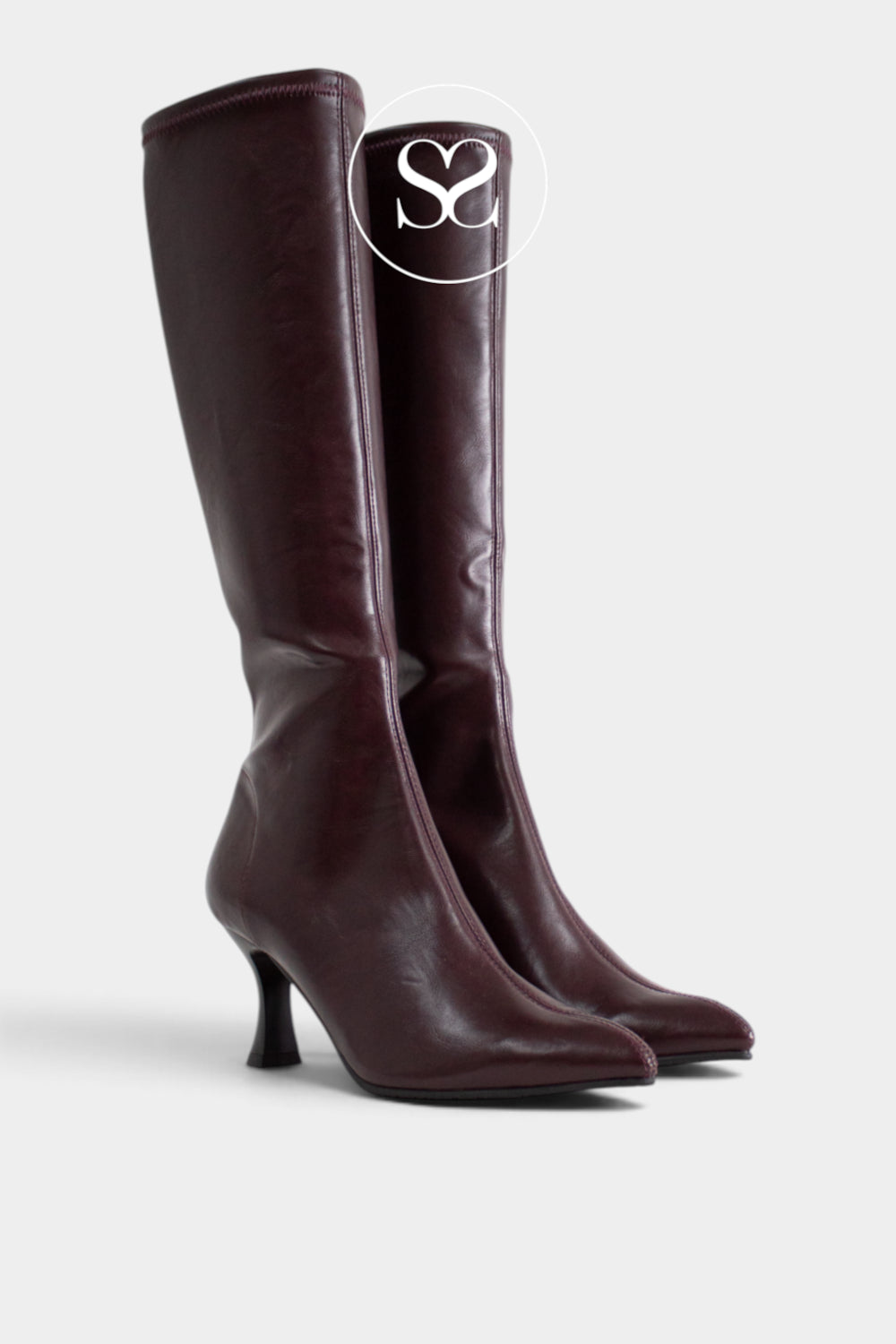 A pair of knee-high pointed-toe boots in a deep burgundy leather, featuring a slim kitten heel and a circular silver 'Sheneil Shoes Ireland' logo overlay on the upper shaft, set against a light grey background.
