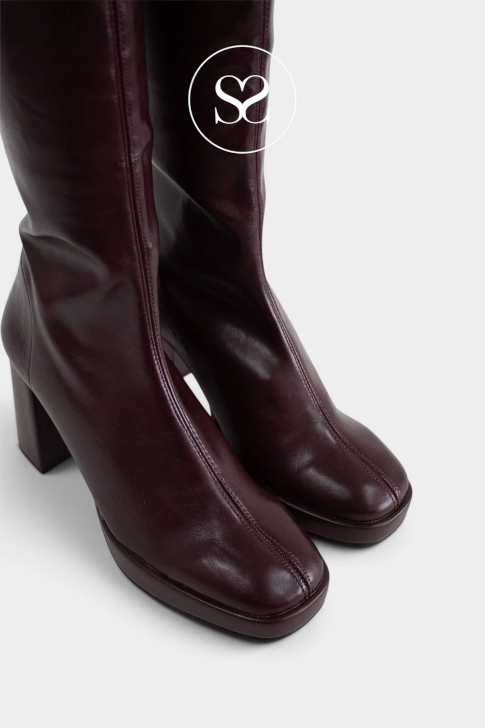 A pair of dark burgundy leather knee-high boots with a chunky block heel and platfrom sole, shown against a light gray background with a white circular 'Sheneil Shoes' logo overlay.