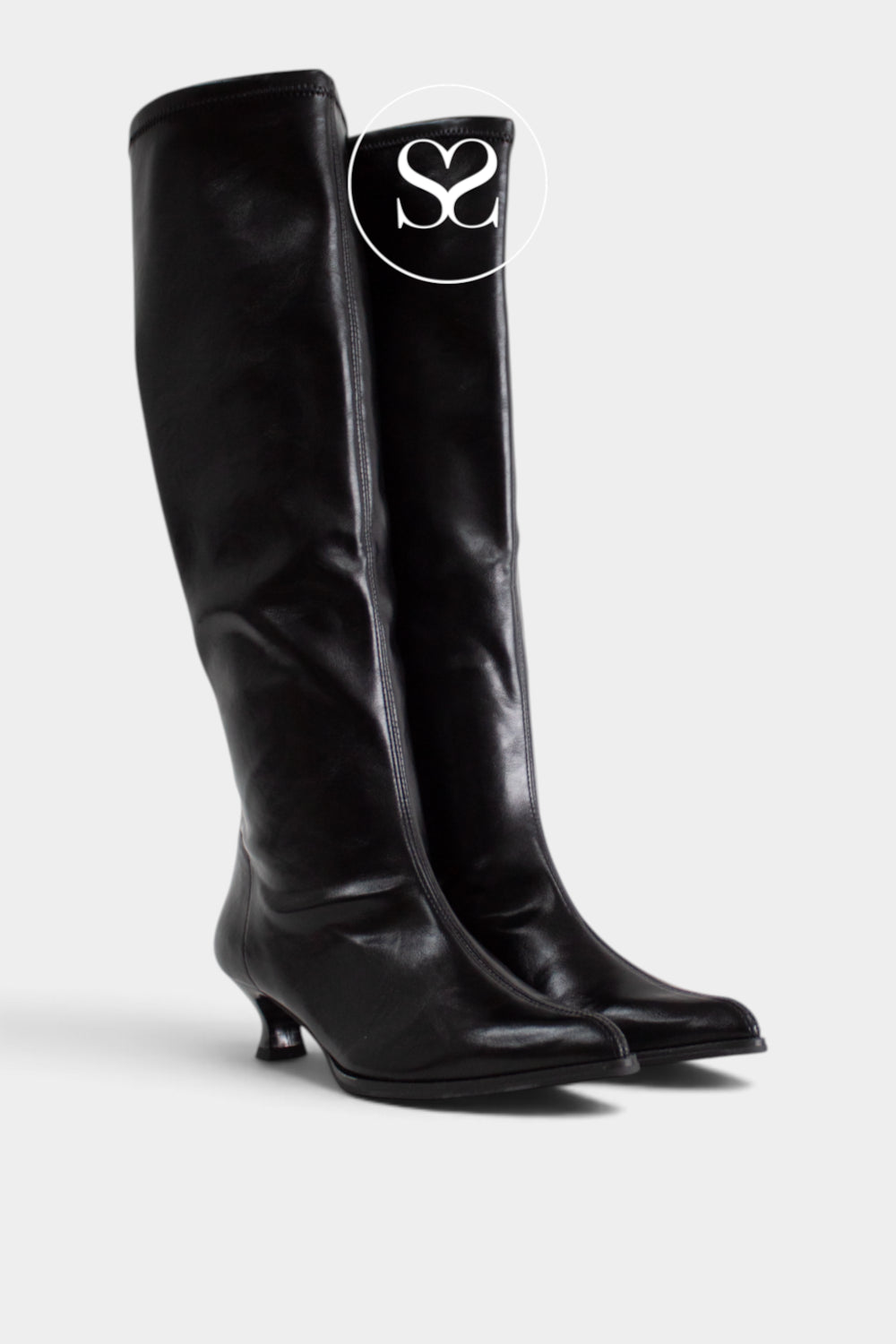 A pair of black leather Miss Elastic knee-high boots with pointed toes and kitten heels, shown on a light gray background. A white circular logo overlay featuring the letters 'Sheneil Shoes' is positioned near the top of the right boot.