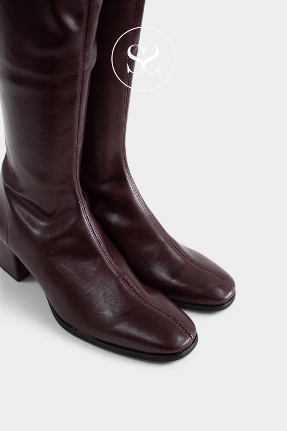 A close-up of a pair of dark burgundy coated knee-high boots with a low block heel and almond toe, shown against a light grey background with a white circular logo watermark.