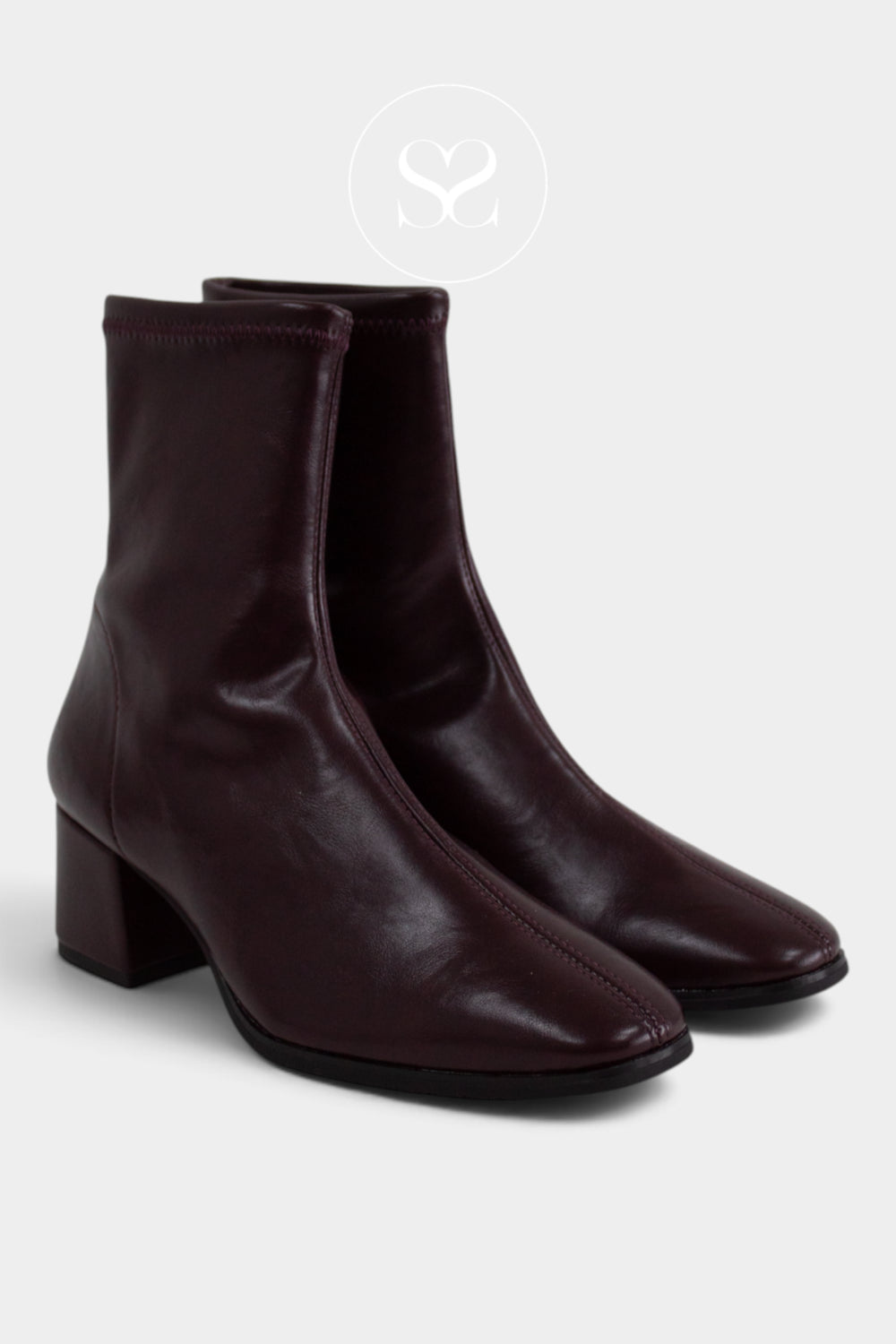 A pair of dark burgundy leather ankle boots with low block heels and almond toes, shown on a light gray background with a faint circular sheneil shoes logo watermark above.
