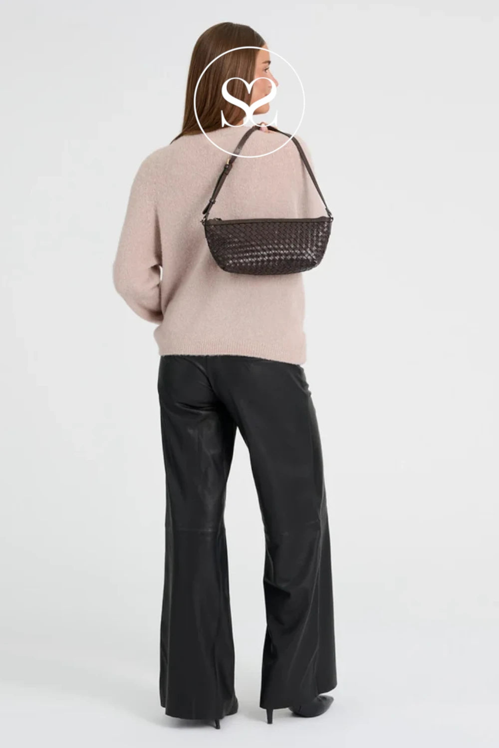 A woman shown from the back wearing a light pink knit sweater, black wide-leg pants, and black heels, while carrying a dark brown woven shoulder bag against a plain light gray studio background.