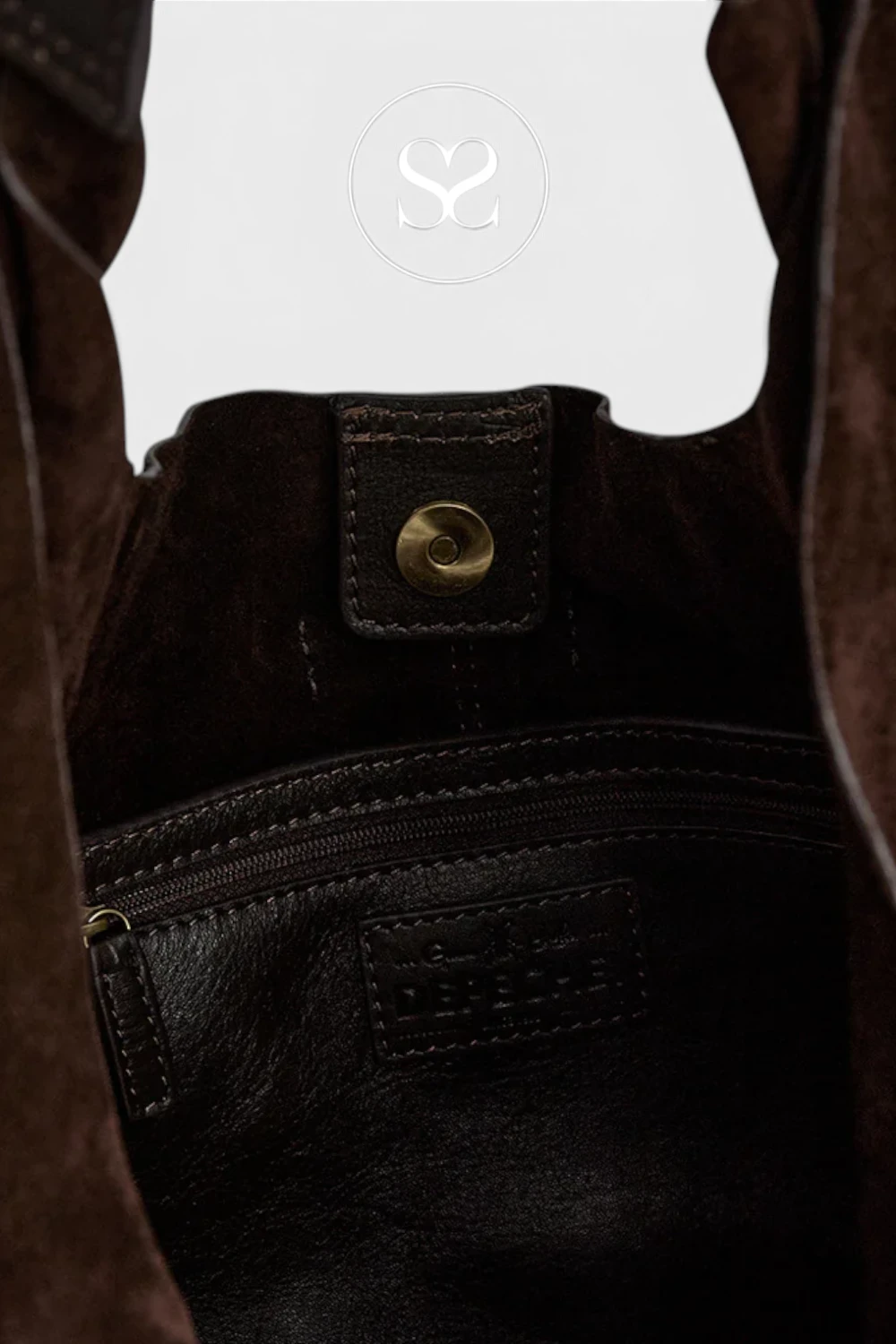 Close-up of the interior of a dark brown leather bag, showing a zipper pocket, a brass snap button, and a small embossed brand patch. A white circular logo overlay with stylized letters is visible near the top.