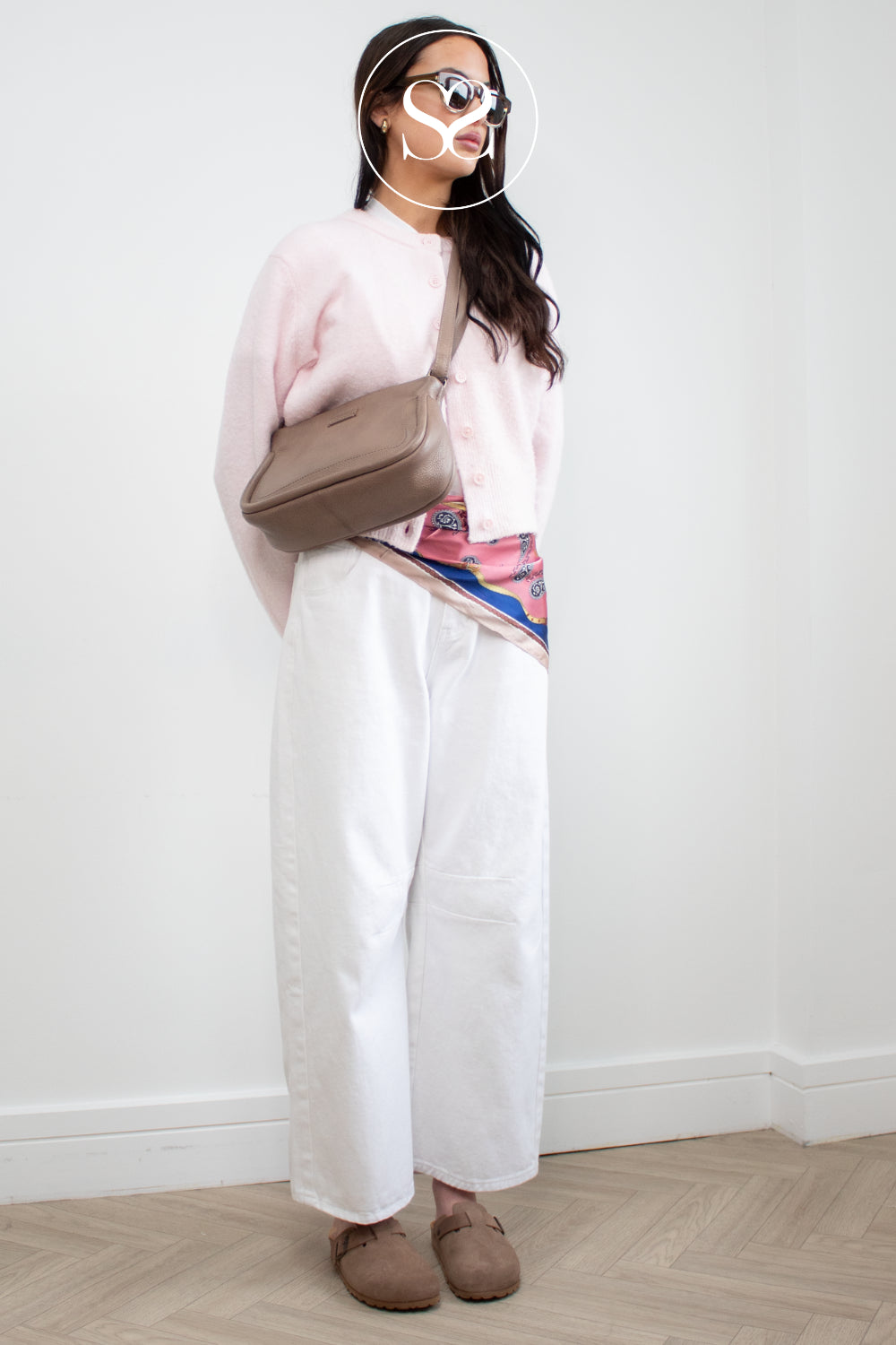 Woman wearing a light pink sweater, white pants, and brown clogs from toni pons, holding a beige Depeche crossbody bag against a white wall.