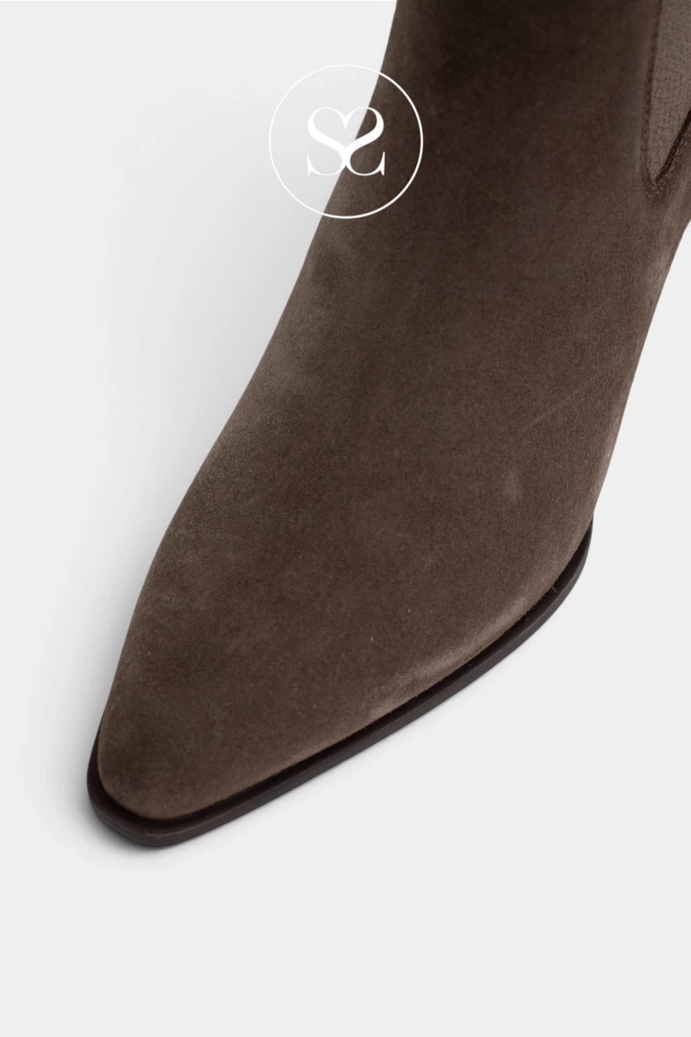 A close-up shot of a Unisa pointed-toe ankle boot in dark brown suede, showing the side profile and a small elastic side panel, on a light gray background with a  Sheneil Shoes watermark logo.