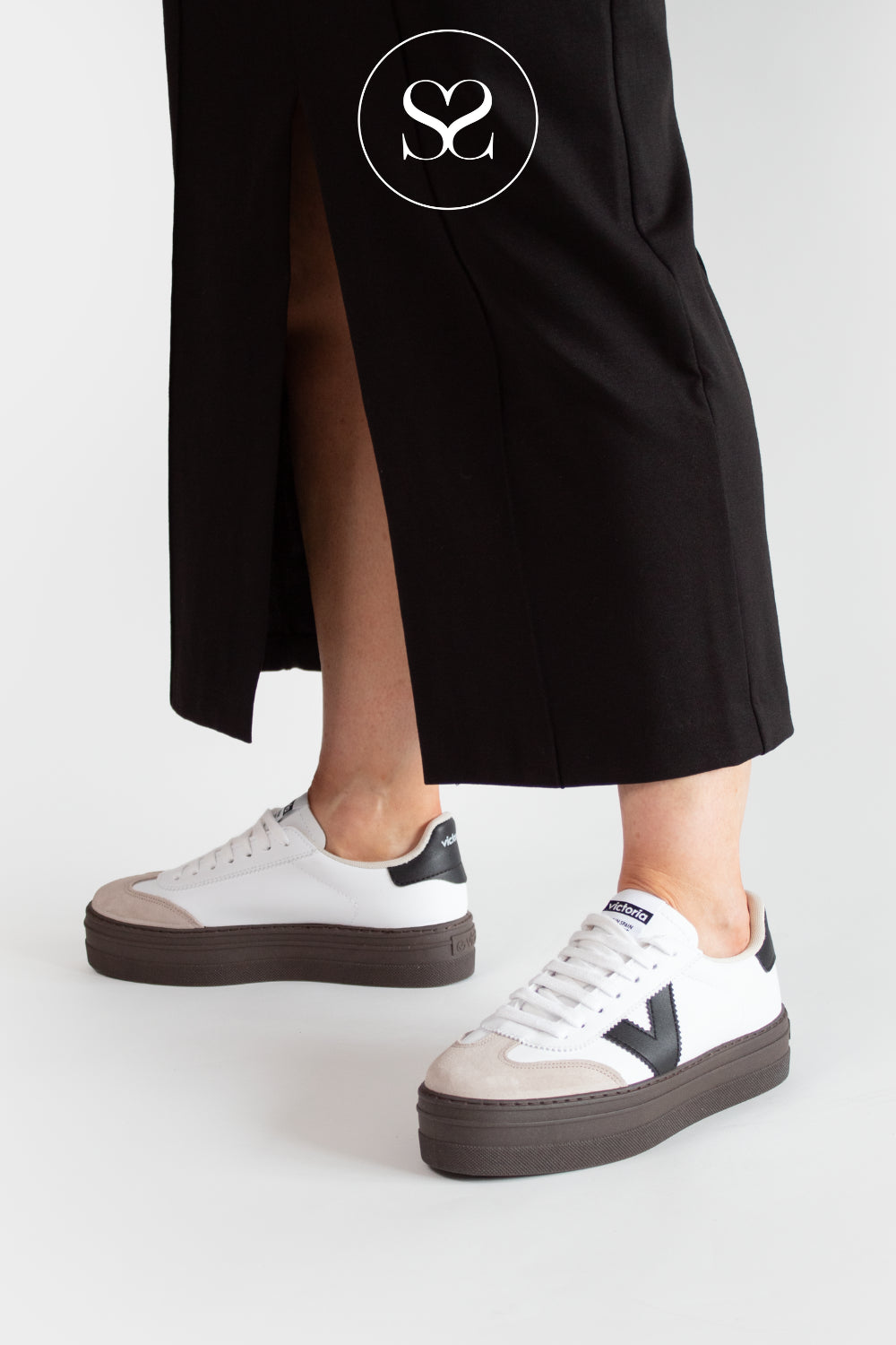 White Victoria platform trainers with brown soles worn by a person in a black outfit on a white background.