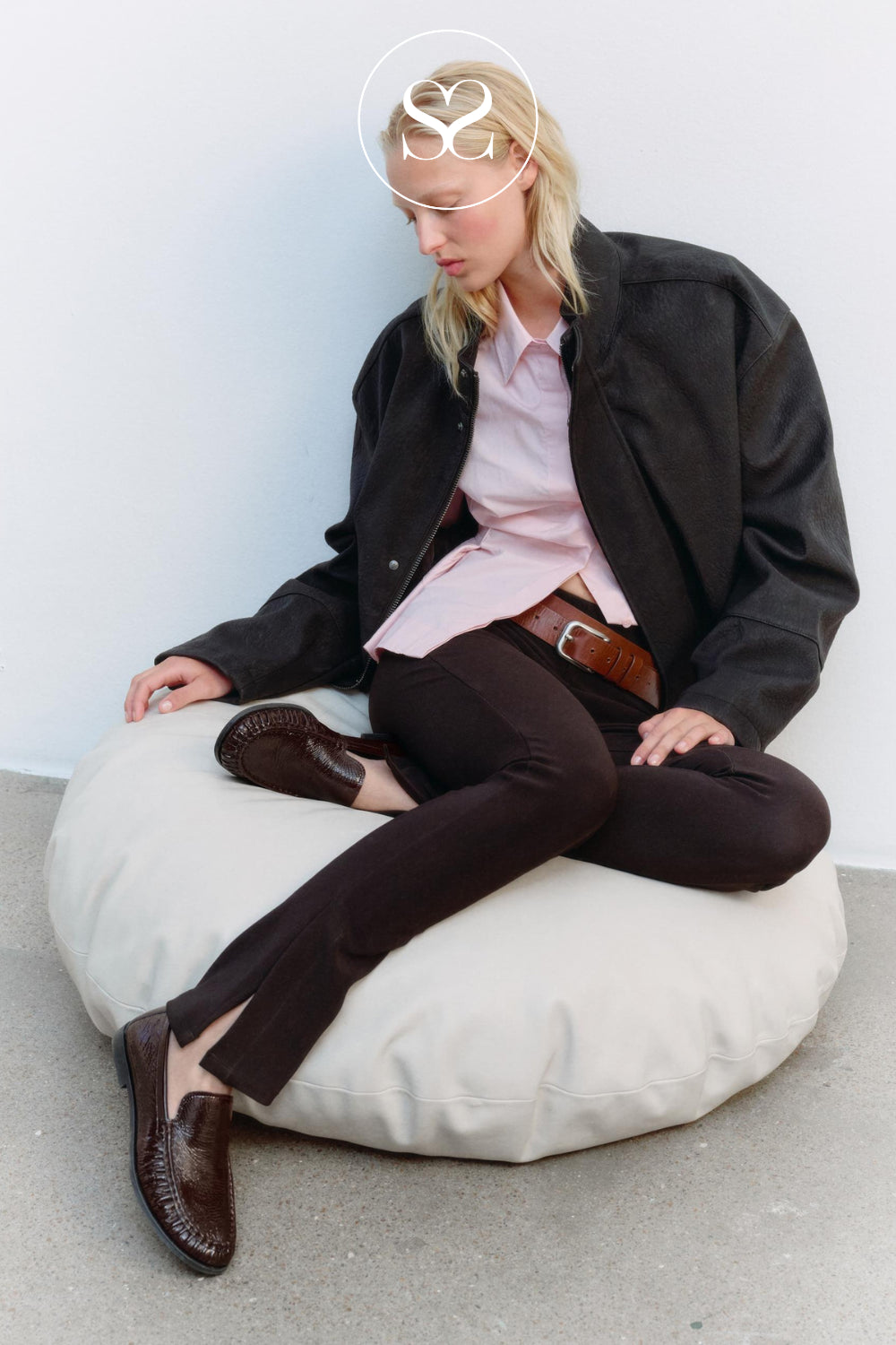 Editorial Images: A model sits on a large cream-colored beanbag chair against a white wall, wearing a black jacket, a light pink button-down shirt, dark brown trousers, and dark brown leather loafers from Vagabond. A white circular logo overlay with 'Shaneil Shoes' is visible near the top center.