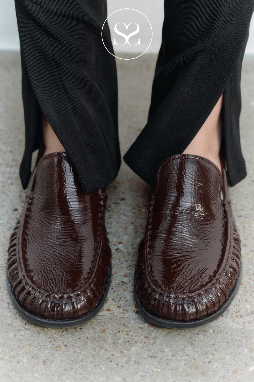 A close-up of a person wearing dark brown textured leather slip-on loafers from Vagabond Shoemakers with black trousers, standing on a light gray concrete floor. A circular Sheneil Shoes logo watermark is visible at the top center.