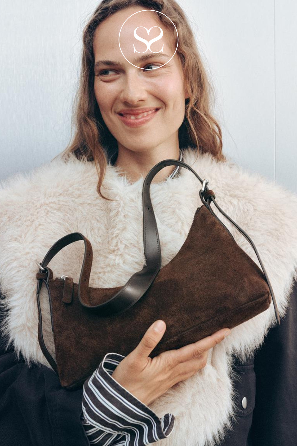 A woman wearing a white faux fur coat holds a brown suede shoulder bag with dark brown leather trim and strap from Vagabond Shoemakers. She is wearing a black jacket over a striped shirt, and a white circular Sheneil Shoes logo watermark is visible on her forehead.