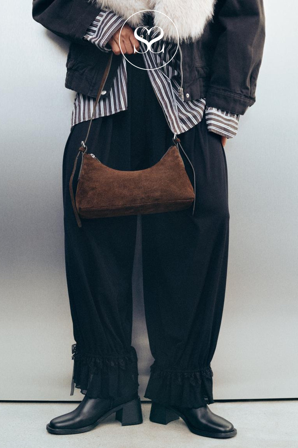 A person wearing a dark chocolate brown suede crescent shoulder bag with a thin strap, styled with black wide-leg ruffled pants, a striped shirt, and black boots against a light gray wall.