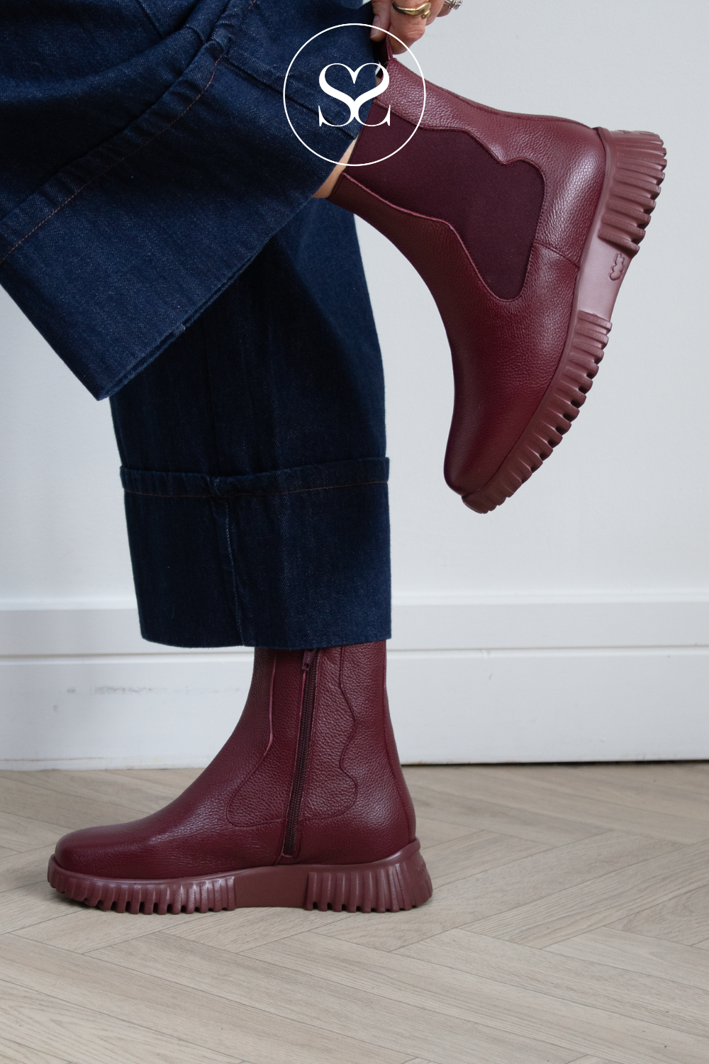 A person wearing Wonders dark burgundy leather Chelsea-style ankle boots with dark blue jeans, shown from the knees down against a white wall and light wood floor. A circular 'Sheneil Shoes' logo watermark is overlaid near the top.