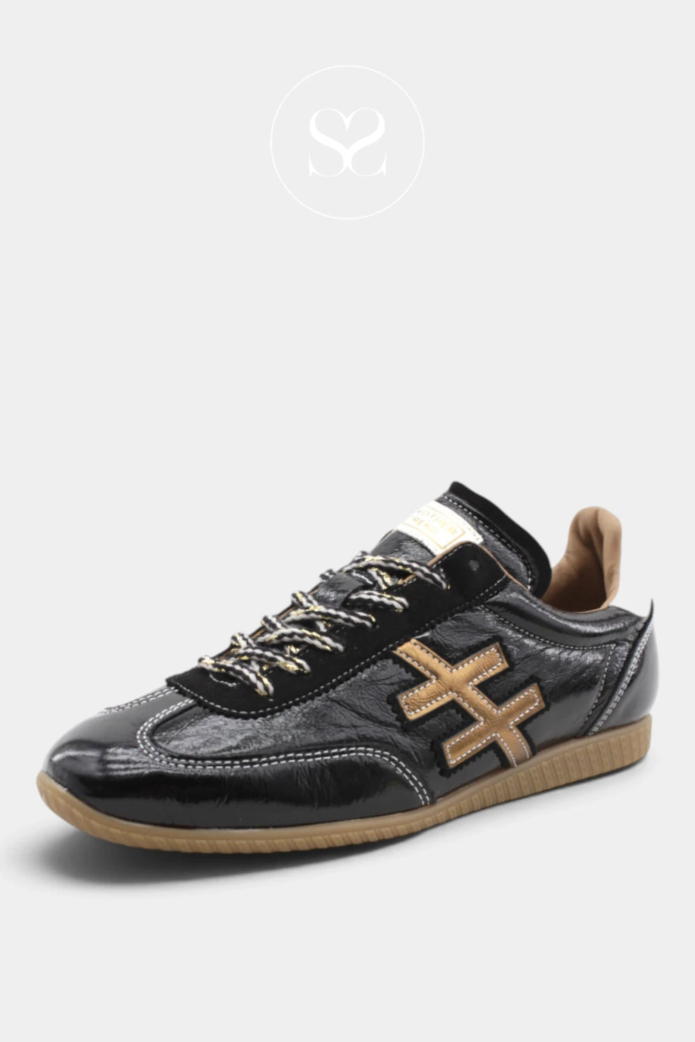 An Another Trend  black leather low-top sneaker with a glossy patent finish, featuring a gold-toned geometric cross logo on the side, gold laces, and a tan rubber sole, shown against a light gray background.
