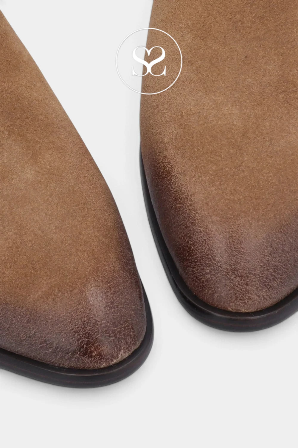 Close-up of a pair of brown suede boots showing the pointed toe and textured leather surface, with a white circular logo watermark at the top center.