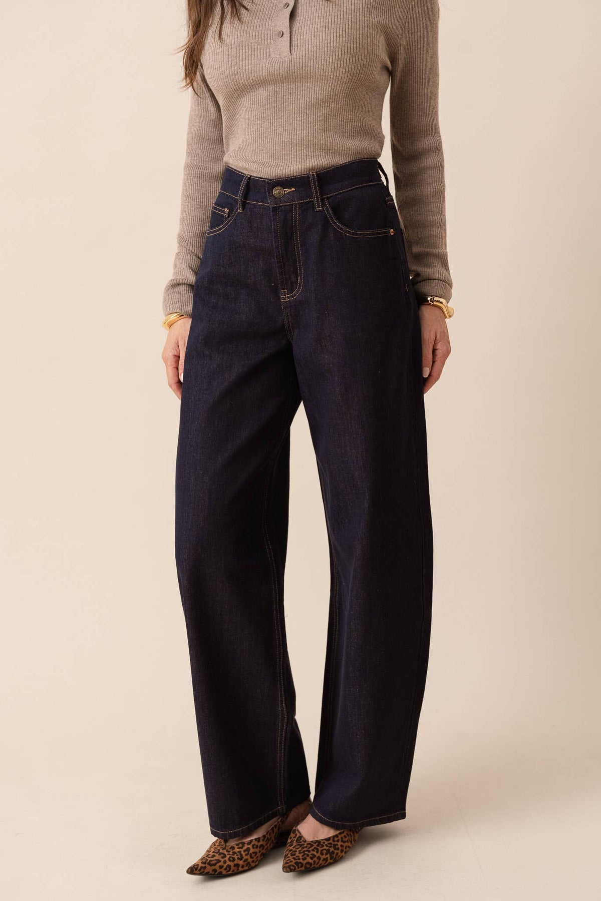 A model wearing dark indigo wide-leg jeans, a beige ribbed knit sweater, and leopard print pointed flats, standing against a solid beige studio background.