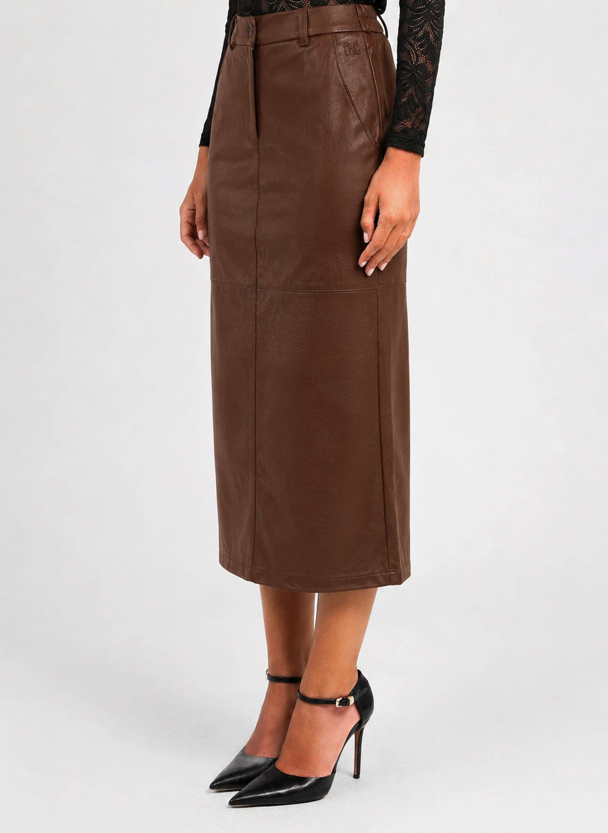 A model wearing a brown faux-leather midi skirt with a matching brown belt, paired with black pointed-toe high heels and a black lace long-sleeve top, shown against a light gray studio background.