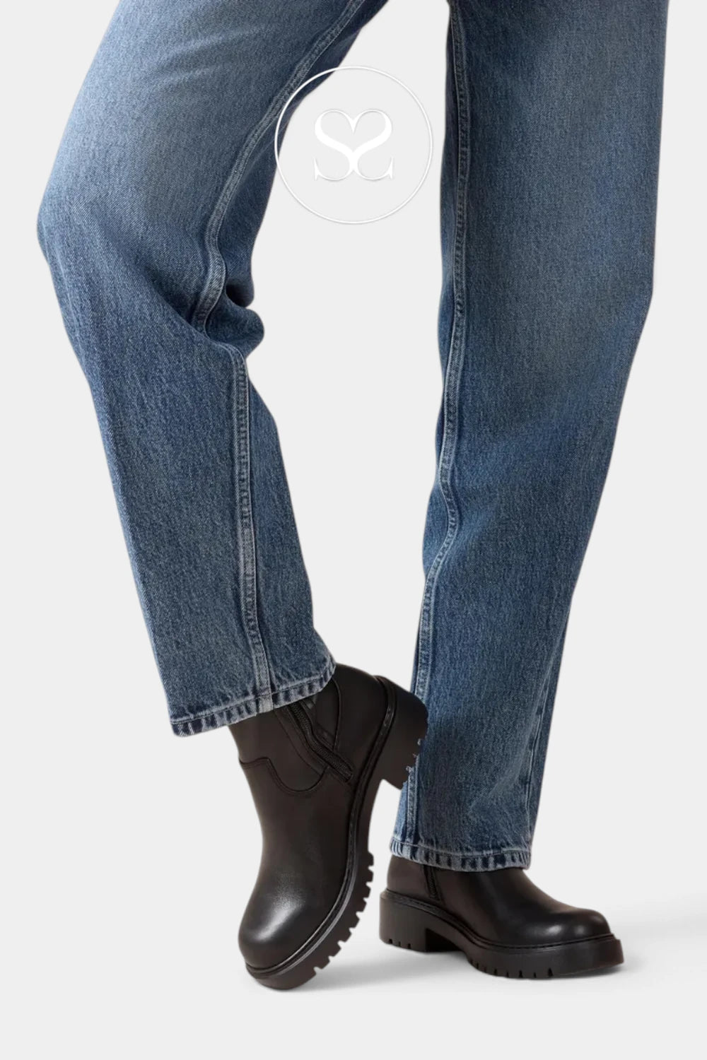 A close-up of a person's legs wearing medium-wash blue jeans and black leather ankle boots with chunky lug soles, shown against a light gray studio background.